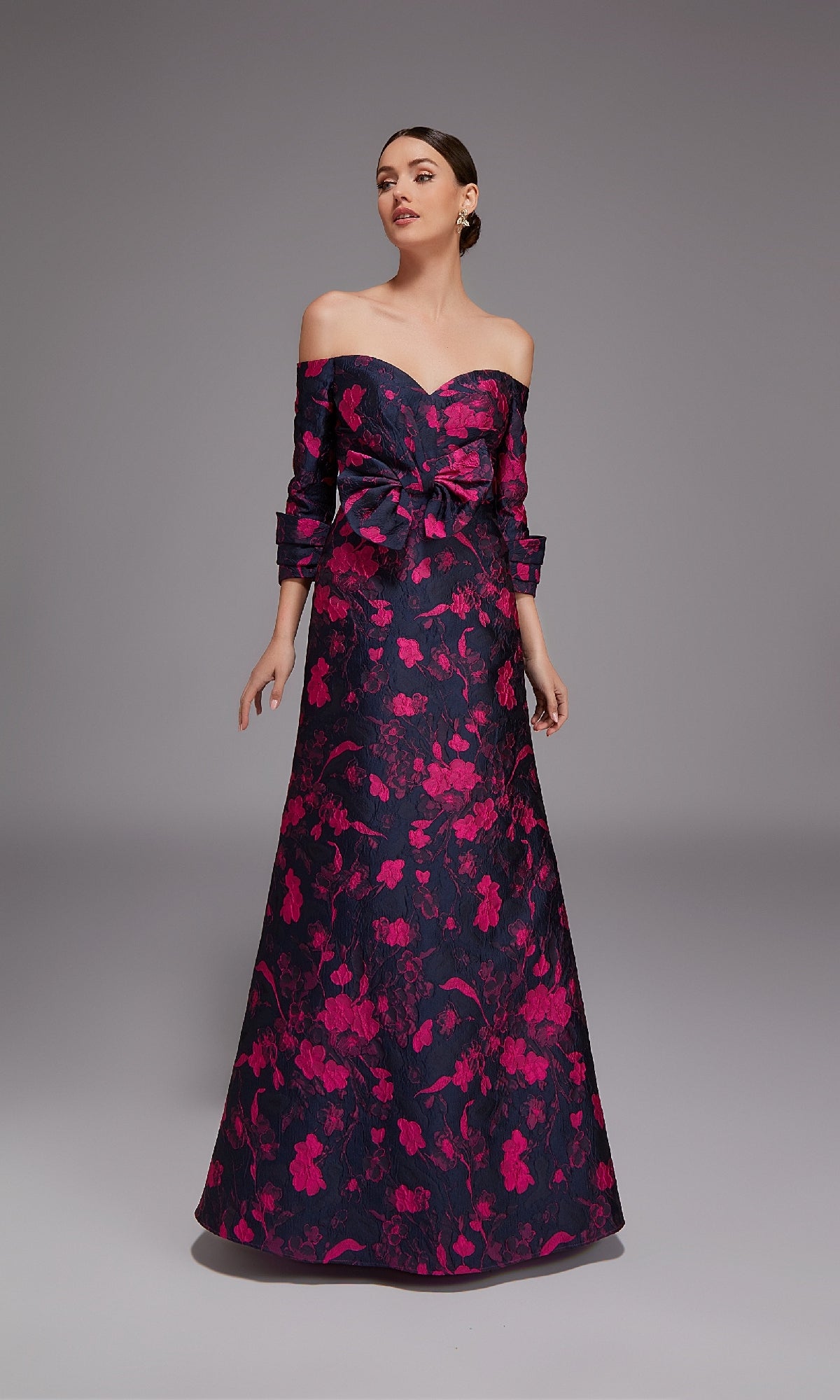 A woman poses against a dark background in the Alyce 27833, a long formal off-the-shoulder dress with an elegant A-line silhouette, bright pink floral pattern, floor-length hem, and a large bow at the waist.